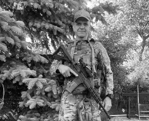 Військовослужбовець у повному спорядженні стоїть просто неба на тлі дерев і тримає перед собою автомат. Чорно-білий кадр передає спокійний момент поза безпосередньою зоною бойових дій. В’ячеслав Шимчук / джерело: Спортивний комітет України, меморіальний проєкт «Янголи спорту»